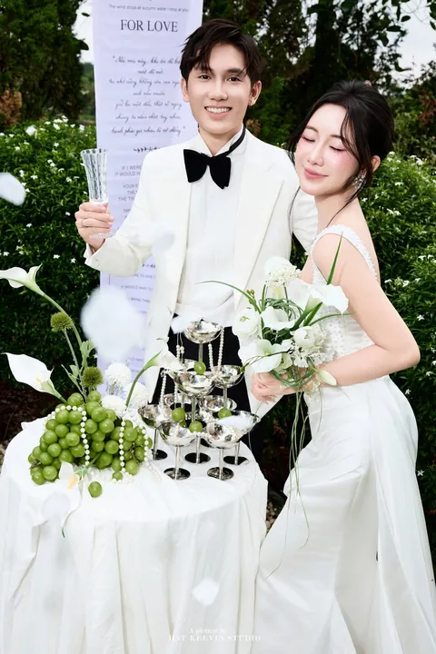 Cô dâu tựa vào chú rể, phía sau là tấm băng lời thề For Love
